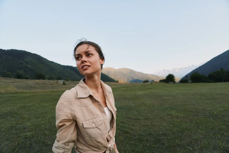 A woman in a beige jacket stands in a field with distant mountains, gazing to the side with a calm, confident gaze, capturing outdoor lifestyle and nature exploration.