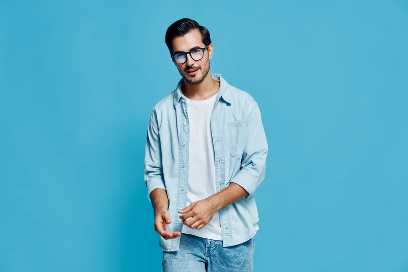 Stylish young smiling man wearing glasses and a denim jacket, standing confidently against a solid blue background. Modern lifestyle and fashion concept.