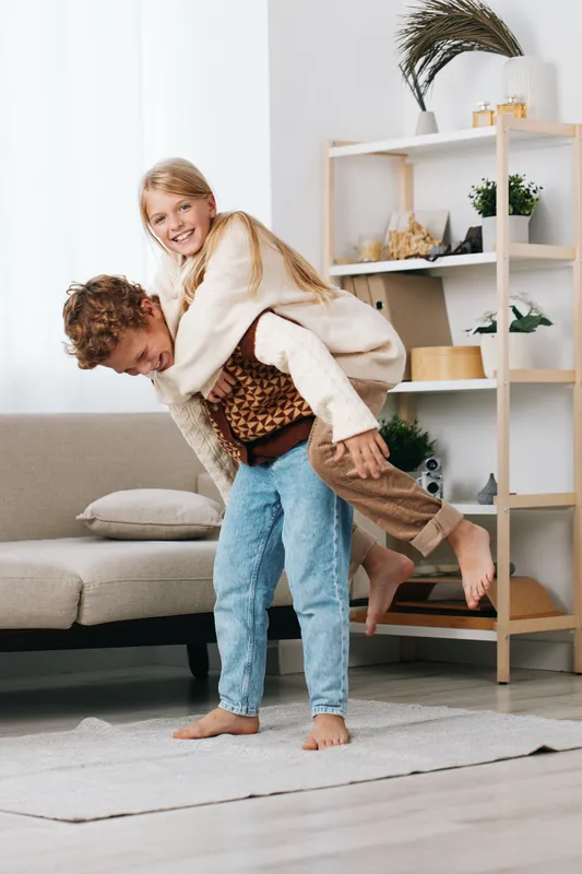 Playful couple in a cozy living room, woman piggybacks on her partner, both smiling. Soft natural light, casual outfits, plush textures, and relaxed, candid moment of affection.