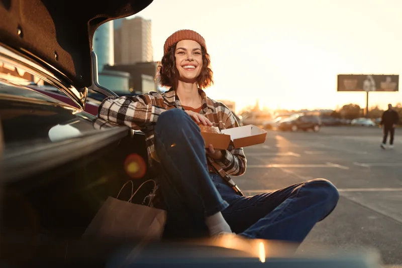 woman car trunk pizza smile parking sunset casual authenticity candid lifestyle golden hour glow mindful living emotional storytelling young woman sits at open trunk eating pizza, smiling in urban