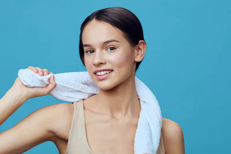 Young woman with a towel on her shoulder smiling, showcasing healthy skin and promoting wellness in a bright blue background, ideal for health and beauty concepts