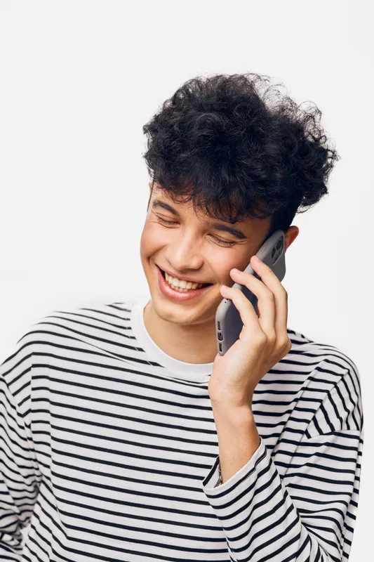 young man smiling, talking on smartphone, casual striped shirt, curly hair, joyful expression, isolated white background, modern lifestyle technology communication