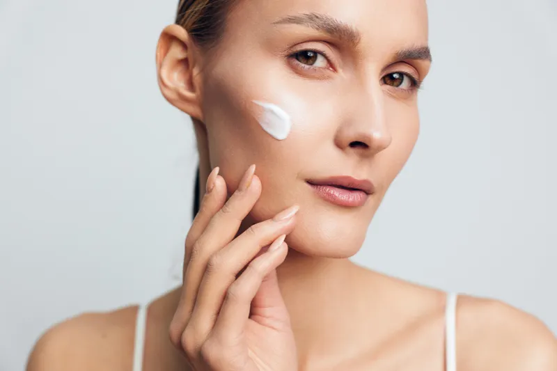 Young woman applies white facial cream on cheek with fingers, has clean skin and natural makeup, skincare routine concept, isolated on light gray background.
