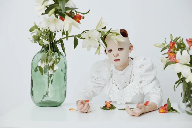 Conceptual art photo featuring a model in a white dress wearing a facial mask, surrounded by flowers, conveying beauty and self care themes