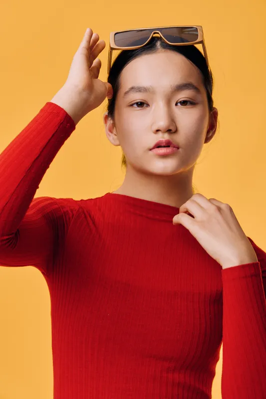 Young woman in red sweater adjusting sunglasses on head against yellow background, posing with calm expression and natural makeup for fashion or lifestyle portrait.