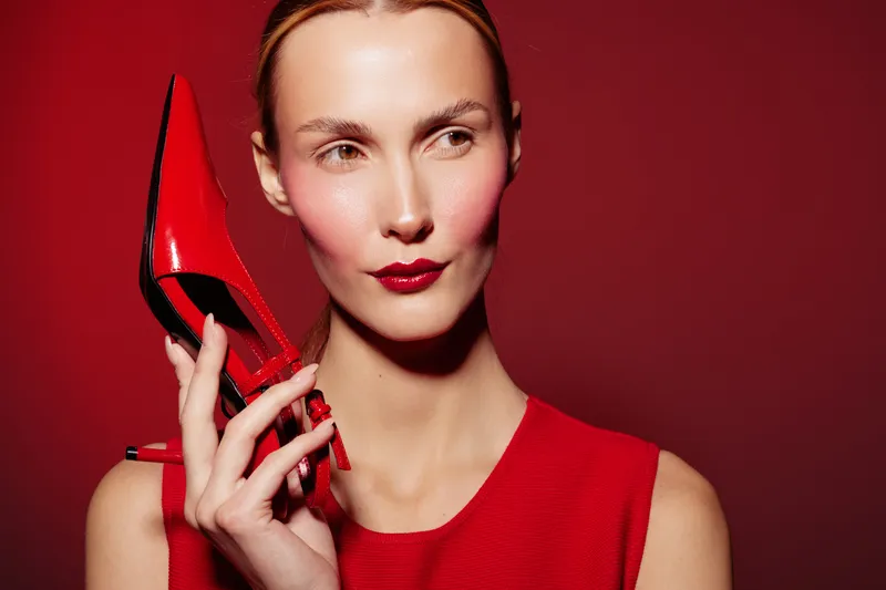 Stylish woman in red dress holds red high heel shoe near face against red background. Fashion concept with bold makeup and elegant accessories in studio lighting.