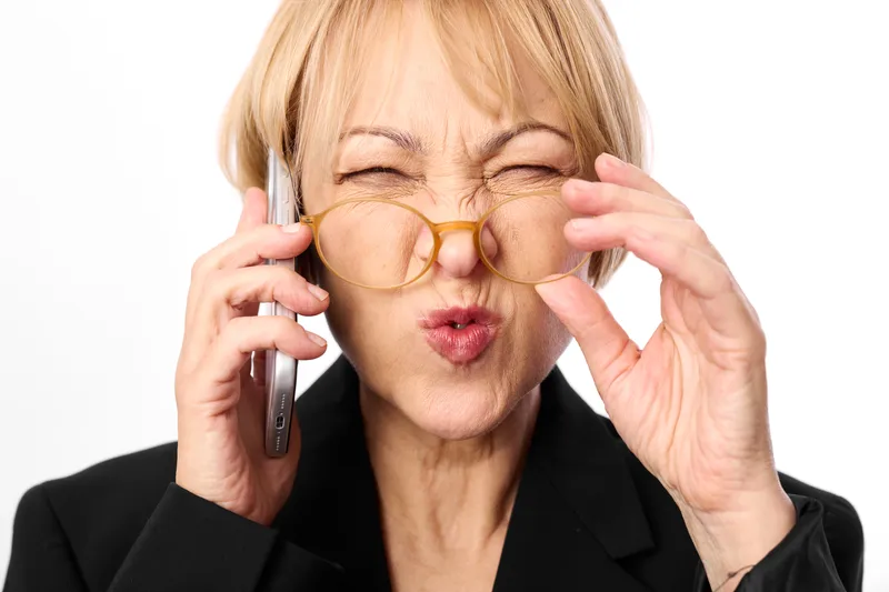 Senior woman in business context holds a phone to her ear and adjusts glasses, showing fatigue and stress in communication. White background, concept for burnout.