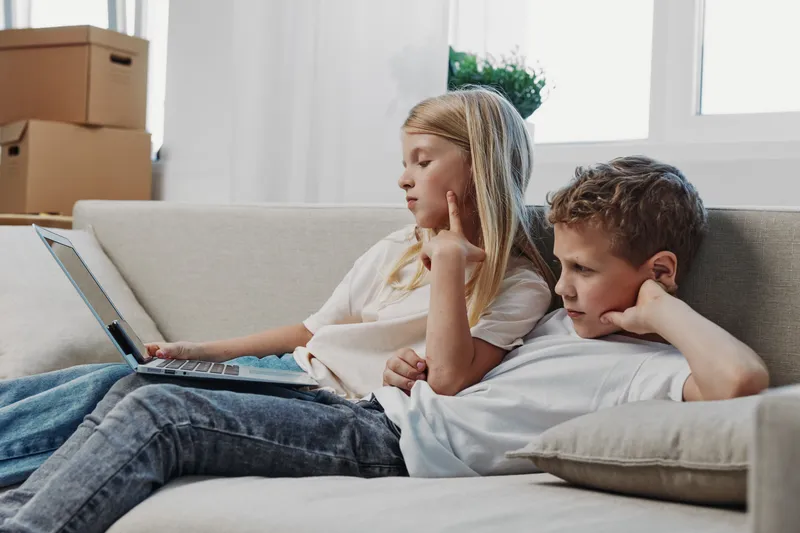 Two children sitting on a sofa using a laptop, focused and relaxed. Bright living room with natural light. Concept of technology, childhood, and casual home environment.