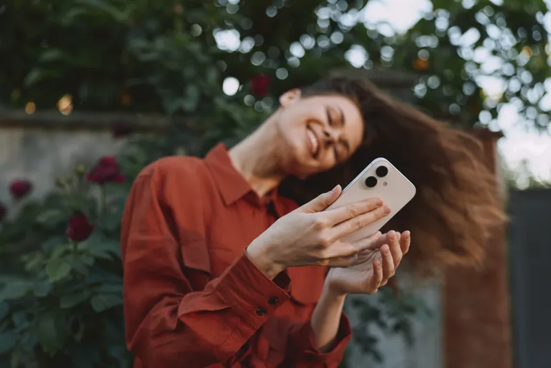 Young woman taking a selfie outdoors, wearing a rust red shirt, hair blown by the wind, bright smile, casual lifestyle portrait in a sunlit garden with natural light and relaxed mood