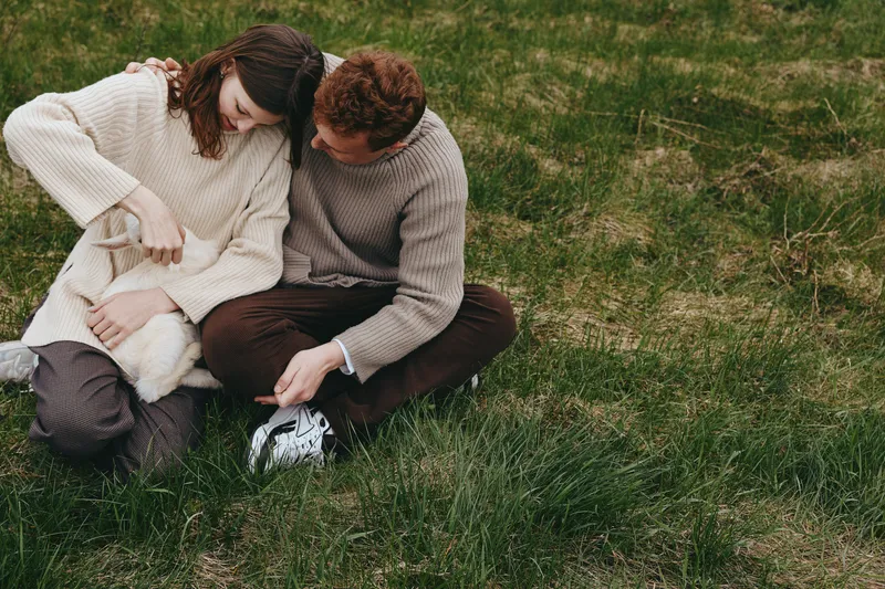 Young couple sits on grass in casual sweaters while caring for a white baby goat in a relaxed outdoor lifestyle scene during autumn.