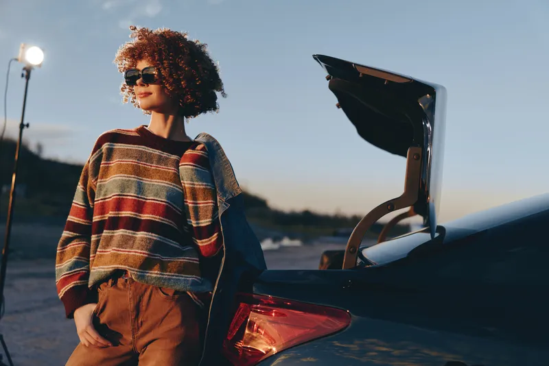 Woman lifestyle outdoors at sunset beside open car trunk, holding a bag, plaid sweater, curly hair, road trip preparation, soft evening fashion light.
