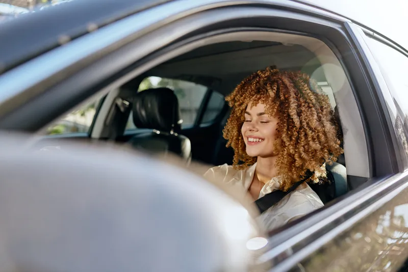 smiling woman driving car with curly hair, seatbelt fastened, daylight, casual shirt, relaxed mood, natural light