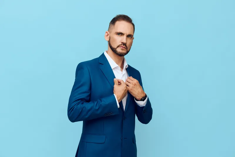 A professional man in a tailored blue suit stands before a solid light blue background, exuding confidence and focus as he adjusts his jacket, signaling calm leadership in a business setting.