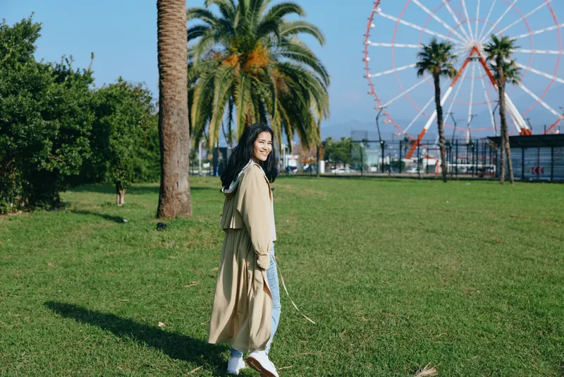 Woman in a light coat walks through a sunlit park with palm trees and green grass, ferris wheel visible in the background, capturing a calm outdoor leisure stroll.