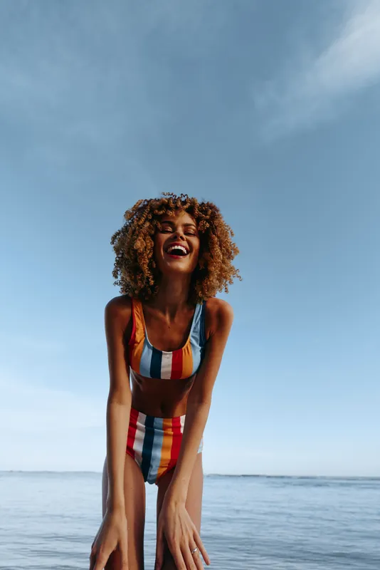 Woman curly hair, ocean swimsuit, smiling outdoors, summer blue sky, standing by sea, sunlit beach, relaxed posture, natural colors, gentle breeze
