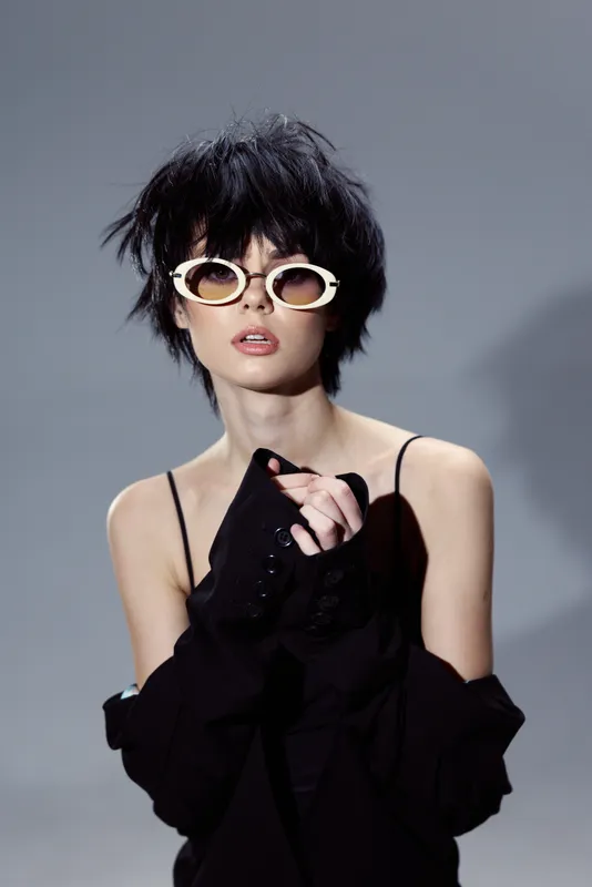 Fashionable woman with short hair wearing white sunglasses and a black off-shoulder outfit in a studio setting