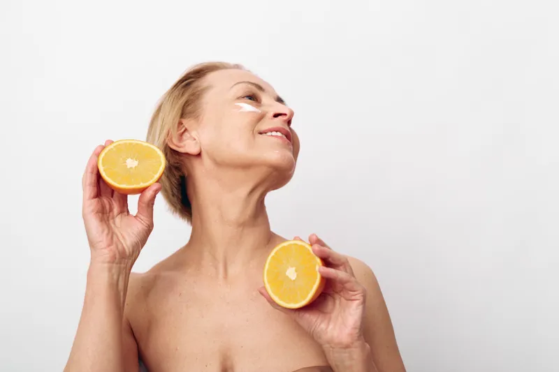 Smiling mature woman with short hair holds two halves of fresh orange near face on white background. Concept of natural skincare, healthy lifestyle, vitamin c beauty and wellness routine.
