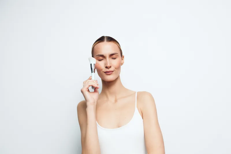 Young woman with clear skin holding facial roller near face, eyes closed, minimal makeup, white tank top, clean background, skincare beauty routine concept.