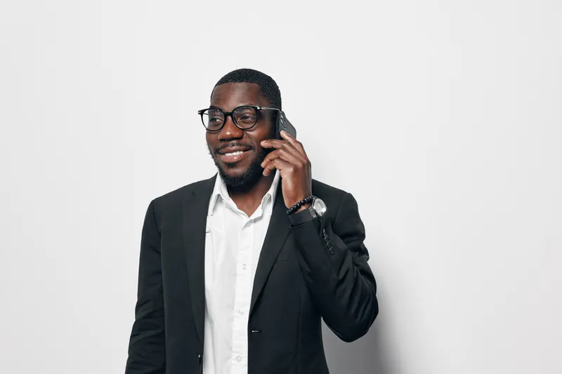 African American man with dark skin wearing glasses and black suit talking on smartphone smiling in studio. Business professional, communication, technology and confident male portrait.
