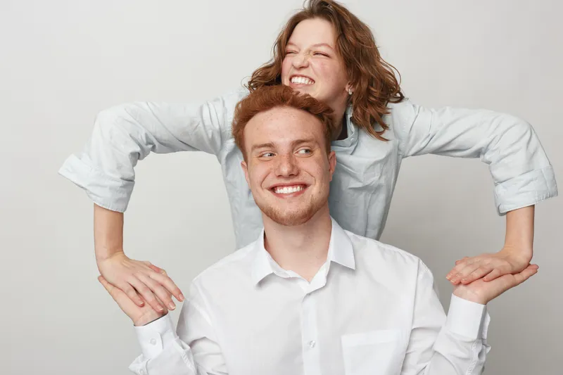 Happy young couple in white shirts playfully posing together on light background. Smiling man and woman express joy and affection in casual studio portrait concept of friendship and love.