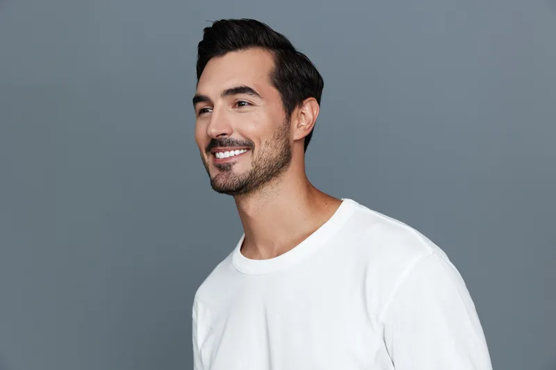 Handsome smiling man in casual white t-shirt looking confidently at the camera with a cheerful expression against a plain grey background. Portrait photography.