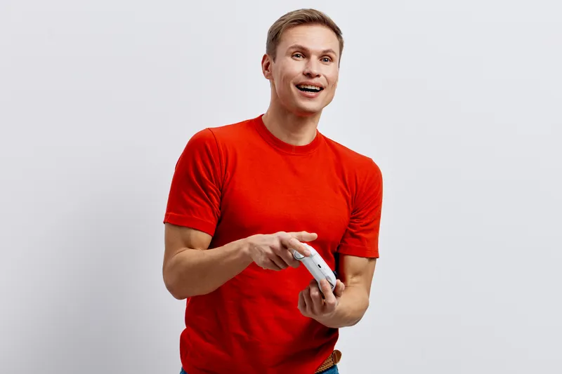 Smiling man in bright red t-shirt holding a remote control, looking excited and happy, standing against plain white background. Casual lifestyle portrait, joyful expression, cheerful mood, studio shot