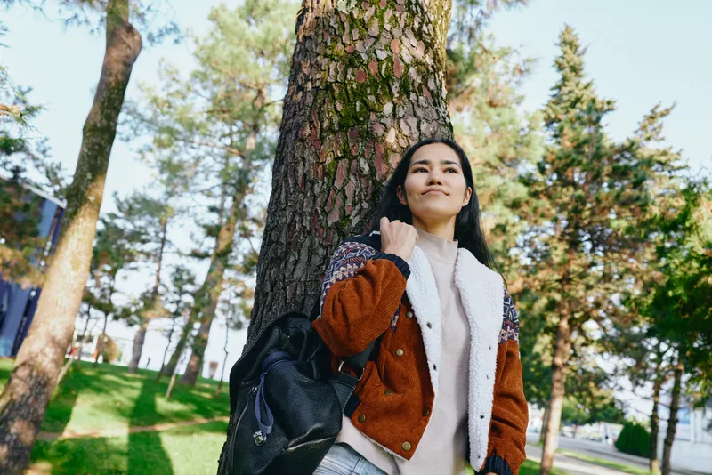 Woman leaning against a large tree in a sunny park, casual jacket and backpack, serene outdoor moment capturing everyday life, nature, and calm.