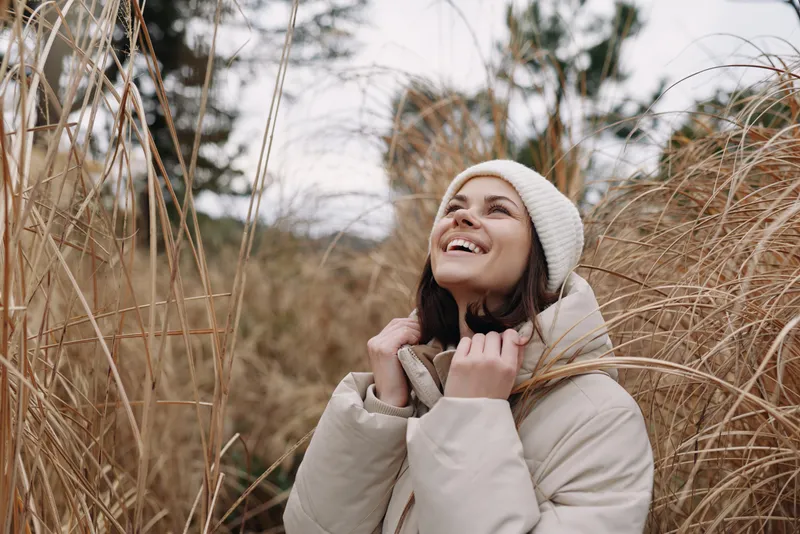 Woman in winter coat and beanie enjoying outdoor nature among tall grasses, smiling up with a warm glow