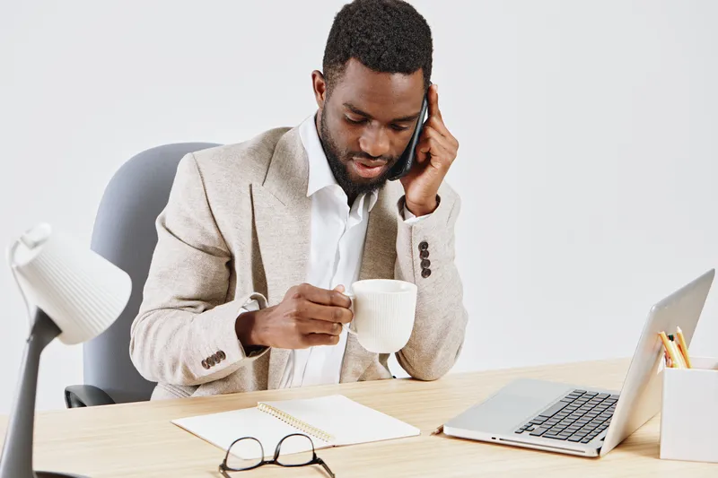 Professional man in beige blazer sitting at desk with laptop and notebook, drinking tea, using mobile phone, in bright office interior, business lifestyle, casual workspace, modern environment, indoor