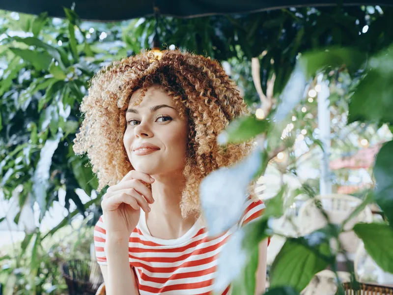 Portrait of a young woman with curly hair resting her chin on her hand among lush green foliage, wearing a red and white striped shirt, bathed in soft natural light.