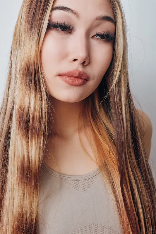 Woman with long straight hair and neutral makeup poses against light background indoors. Female model with natural expression wearing sleeveless top in closeup portrait for beauty or fashion.