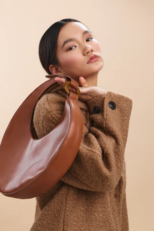 asian woman wearing a tan coat holds a curved brown bag over her shoulder in a studio portrait, exuding chic confidence and refined elegance with a minimal background.