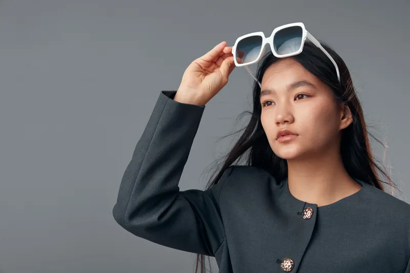 Young woman wearing dark blazer adjusting white sunglasses on head looking away with long black hair isolated on gray background in studio portrait fashion and style concept.