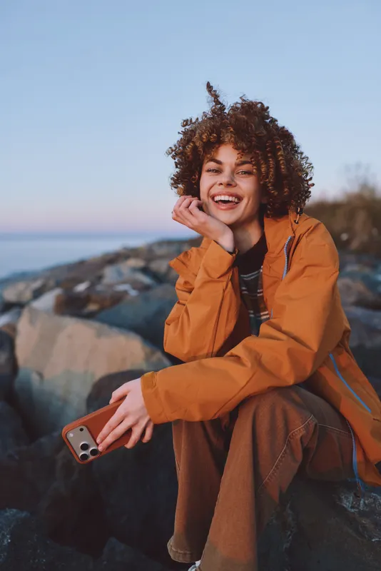 Smiling woman with curly hair in orange jacket sitting on coastal rocks, chin resting on her hand, holding a smartphone, outdoor lifestyle travel mood, soft sunset light, calm