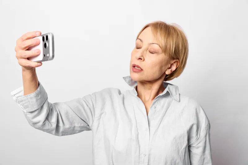 Woman selfie smartphone portrait smile technology communication, white shirt background, short hair holds phone up, candid moment, modern indoor lighting