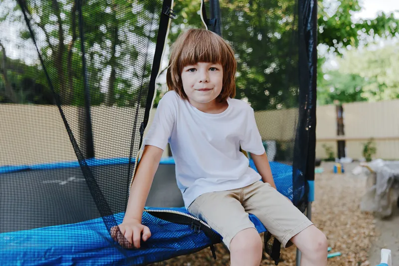 Young boy enjoying time on a trampoline, wearing a casual white t-shirt and shorts, surrounded by greenery in a playful outdoor setting.