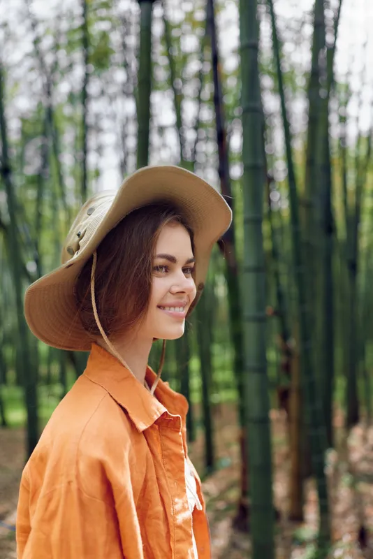 Woman portrait smiling in bamboo forest wearing a wide-brim hat and orange shirt, side profile travel lifestyle, nature outdoor scene with soft natural light and relaxed expression.