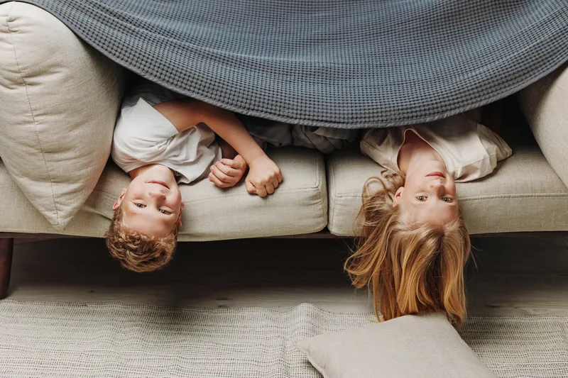 Two happy children lying upside down on a beige couch covered with a gray blanket. Casual home scene with natural light, playful mood, and cozy living room atmosphere.