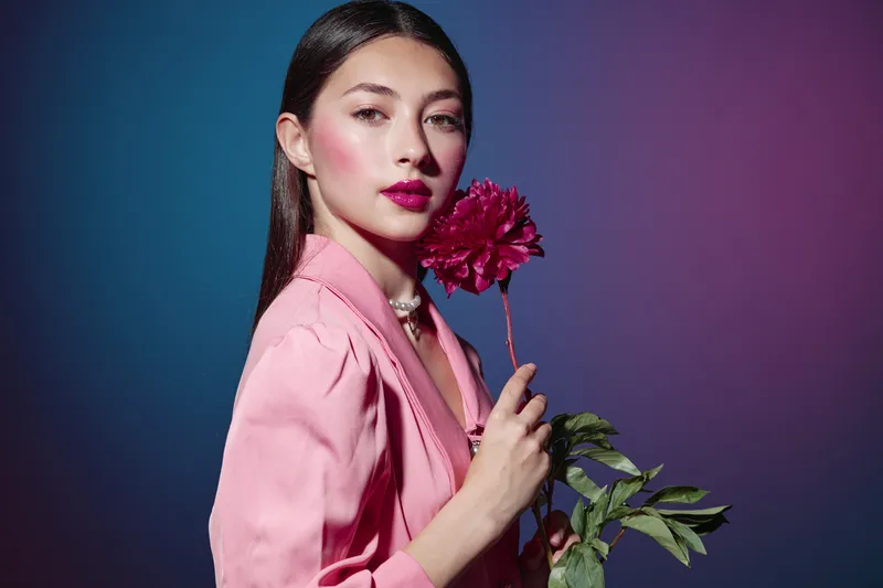 Young woman with pink makeup and red lips holding red flower wears stylish pink blazer against colorful gradient background. Concept of beauty fashion creative portrait.