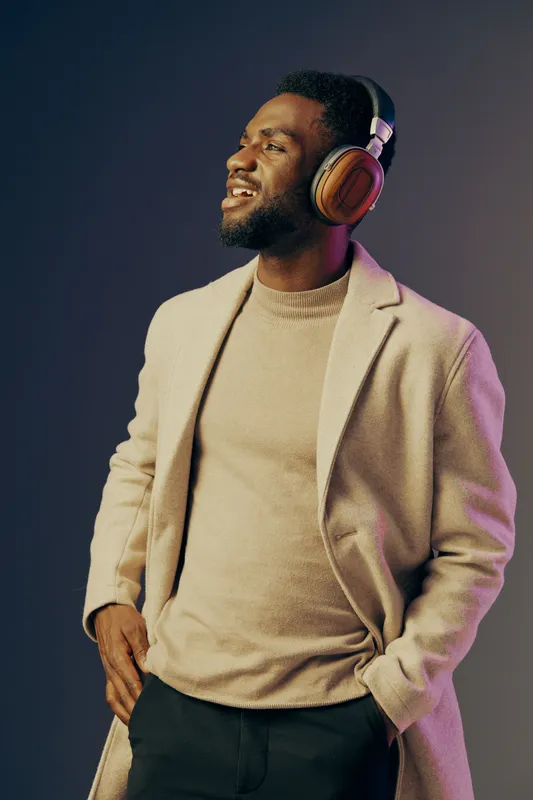 Smiling Black man in stylish headphone, wearing beige coat and sweater, set against a gradient background, conveying happiness and confidence