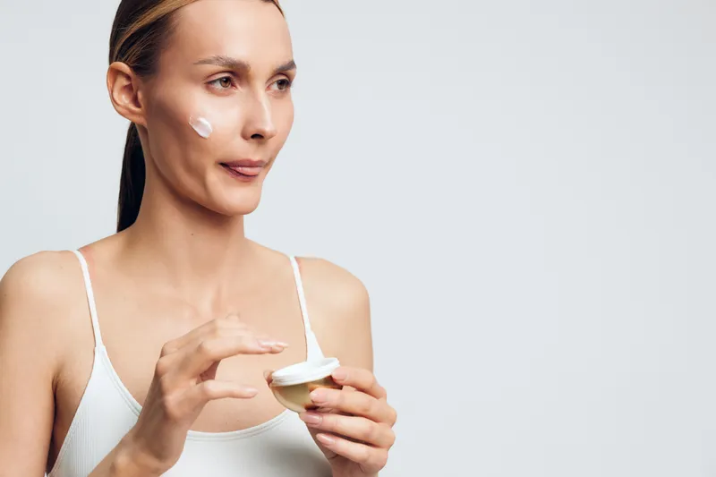 A woman applies moisturizer to her face, illustrating skincare focus and selfcare routine in a bright, minimal setting, with emphasis on hydration, healthy complexion, and glowing skin.
