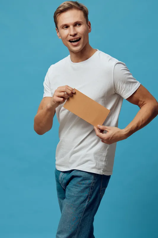 Happy young man with casual white t-shirt holding a blank cardboard sign, standing against a solid light blue background, looking cheerful and engaging for promotional or advertising use. People lifes