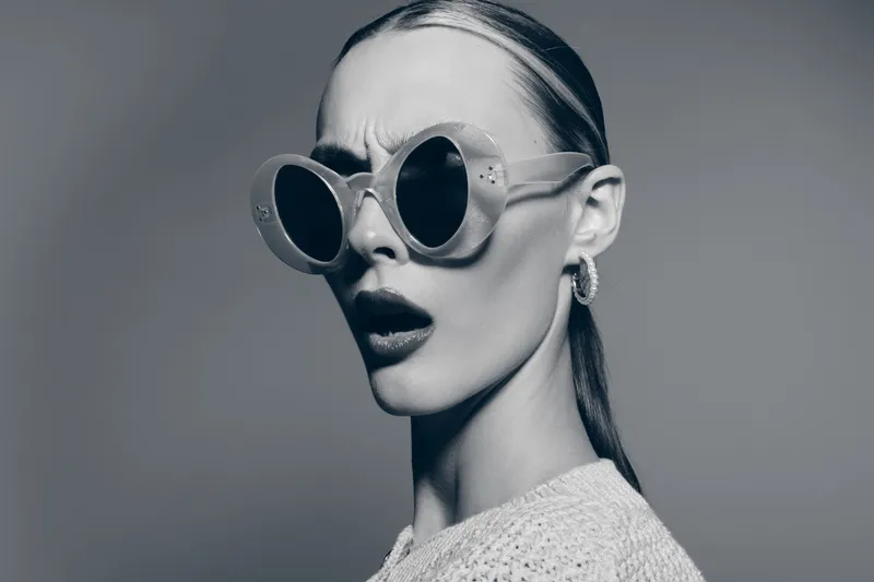 Young Caucasian woman with pale skin wearing round sunglasses and knitted sweater looks surprised, monochrome studio portrait.