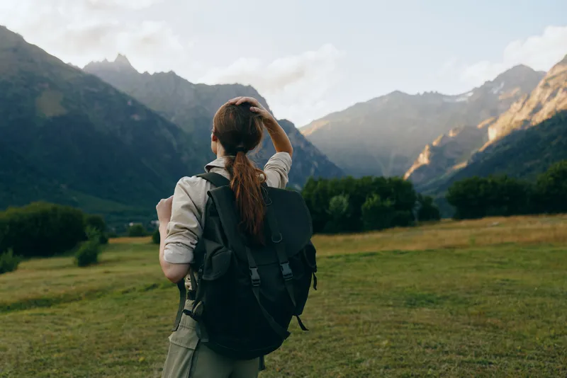 Young woman with a backpack surveys a green valley as rugged mountains rise in the distance under a clear sky, conveying adventure and quiet anticipation.