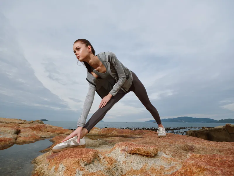Young woman practicing yoga on rocky beach during overcast day, showcasing flexible pose, athletic wear, and serene ocean backdrop