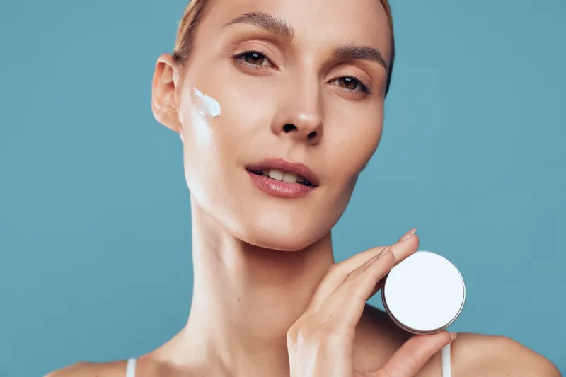 Young woman with fair skin applies moisturizer on her cheek while holding a cosmetic jar against a plain blue background in a skincare routine scene.