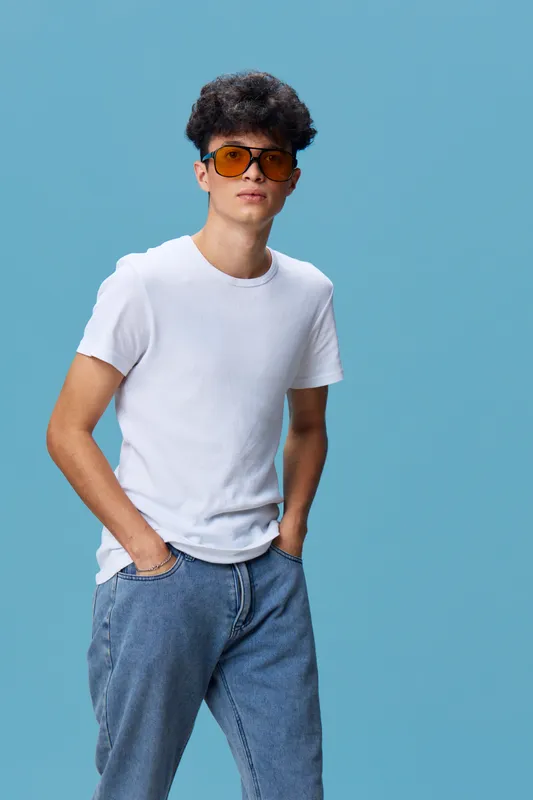 young man wearing sunglasses and white t-shirt standing confidently with hands in pockets against blue background, casual denim jeans, modern fashion look