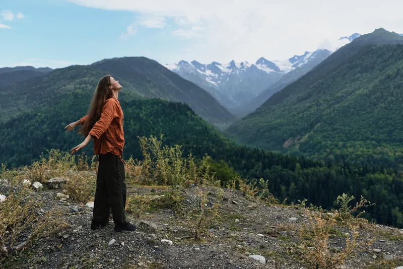 A lone woman stands on a rocky overlook, arms outstretched, admiring a dramatic mountain valley with snow capped peaks beneath a clear blue sky.