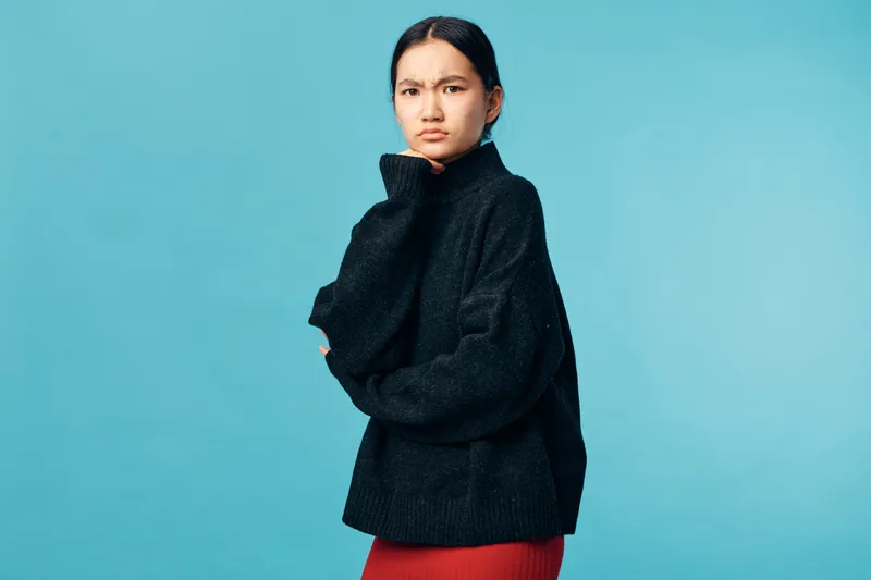Woman in black sweater and red skirt standing against blue background looking thoughtful with hand on chin, casual style and serious expression in studio portrait.