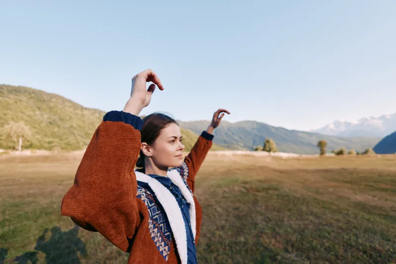 woman in nature with mountains and meadow background, autumn outdoor portrait wearing warm jacket, arms raised in joyful pose, serene landscape and soft daylight for travel lifestyle
